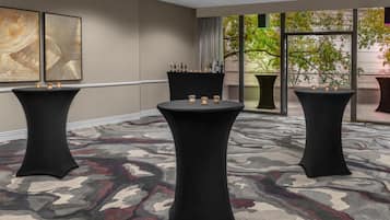 Un salón de conferencias con tres mesas redondas, cada una cubierta con un mantel negro y velas. La sala tiene una alfombra estampada, ventanas amplias con vistas a árboles y arte abstracto en las paredes.