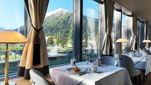 A dining area with tables set for two, large windows offering a view of a mountain and lake, and modern lamps on each table.