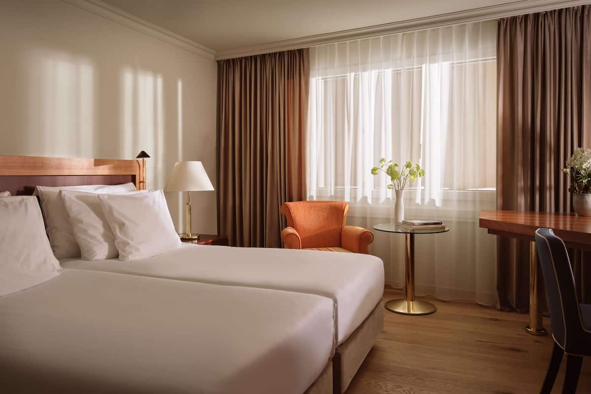 A hotel room with two beds, a wooden headboard, a small table with a lamp, a chair, and a view of the outdoors through large windows with curtains.