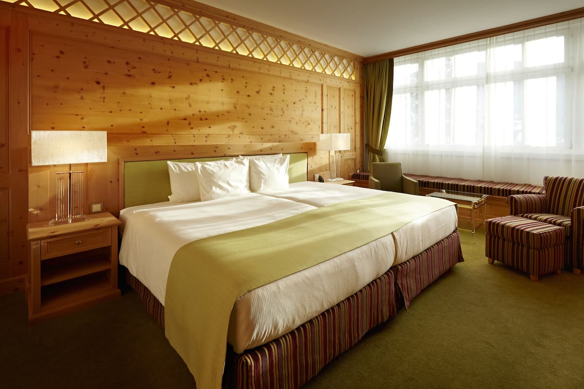 A hotel room with a large bed, two bedside tables with lamps, a chair, a bench, and a large window with curtains.
