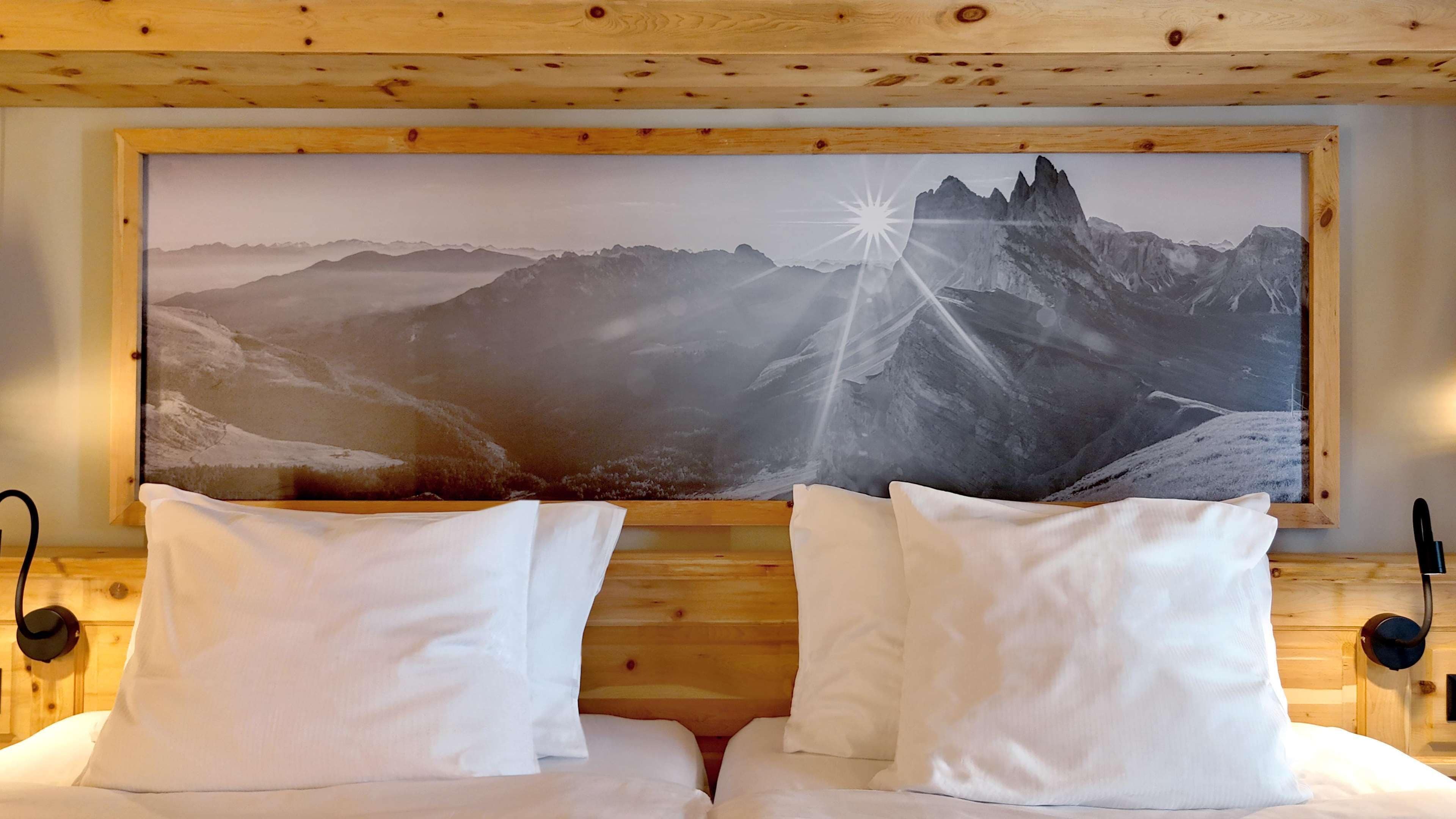 A bedroom with a large mountain landscape painting above the bed, wooden headboard, white bedding, and a pair of black bedside lamps.
