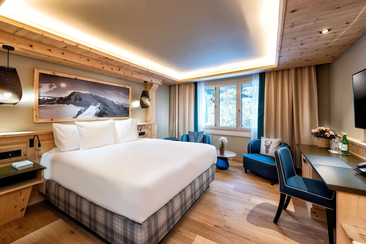 A modern hotel room with a large bed, a desk with a chair, a television, and a window with curtains.