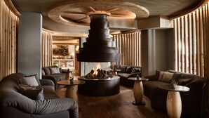 A modern lounge area with a central fireplace, sofas, and round tables.