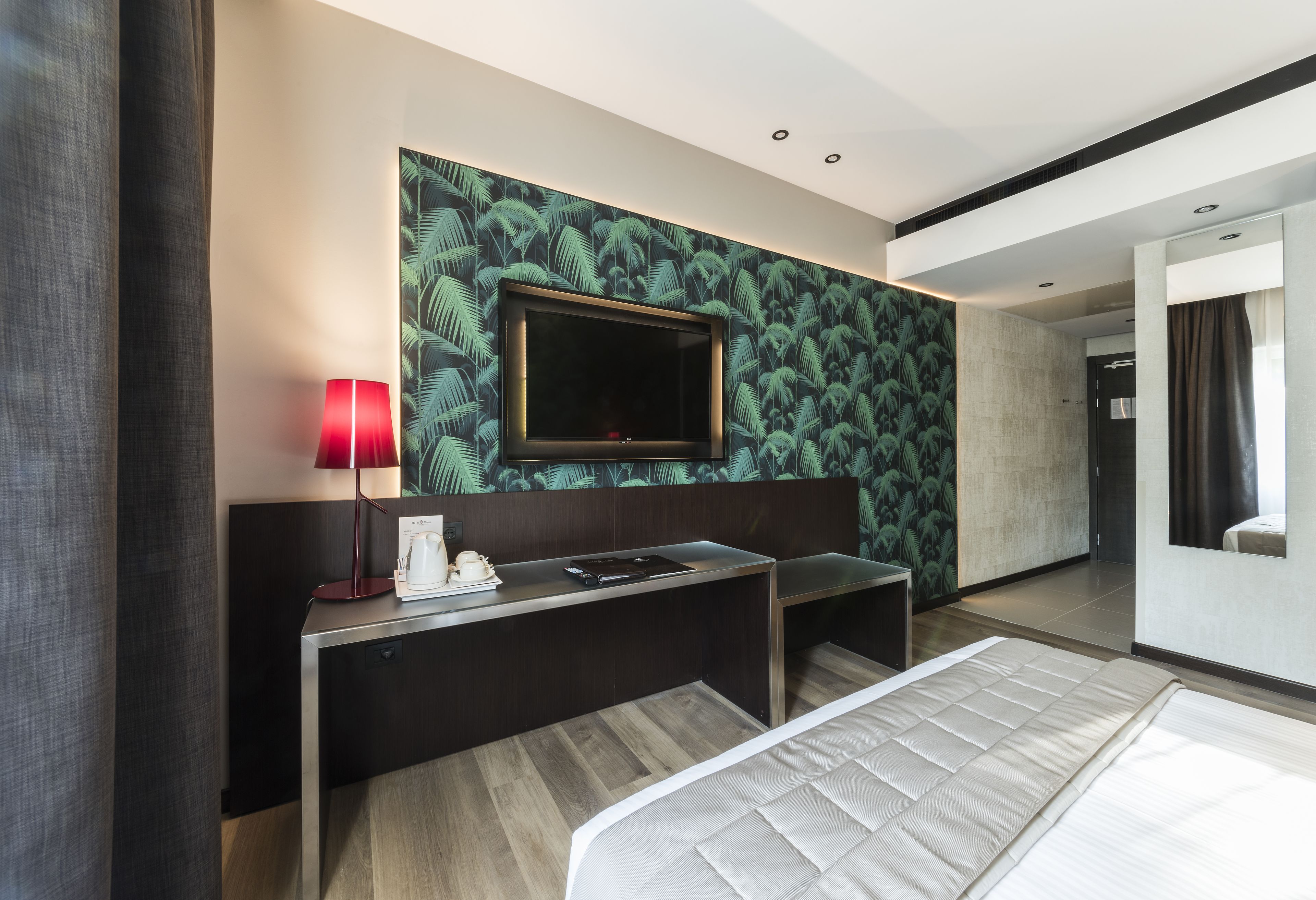 A modern hotel room with a large bed, a desk with a red lamp, a flat-screen TV, and a tropical wallpaper.