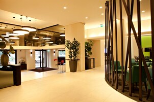 Een moderne lobby met een receptiebalie, potplanten en een zithoek met groene stoelen.