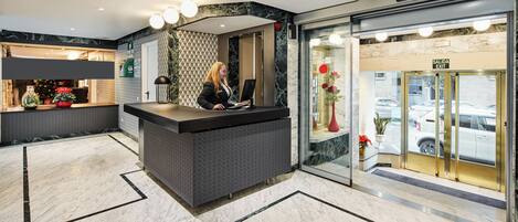 Zona de recepción de un hotel con suelo de mármol, mostrador de recepción y puerta de cristal que da al exterior.