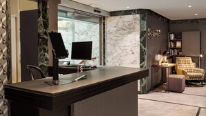 A reception desk with a flat-screen monitor, a keyboard, and a mouse. There is a bookshelf with books and a globe. A plaid armchair and a small ottoman are visible. - Sercotel Togumar (Madrid)