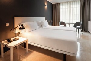 Hotellihuone, jossa on suuri sänky, yöpöytä lampun kanssa ja tuoli ikkunan lähellä, ja ikkunassa on verhot.