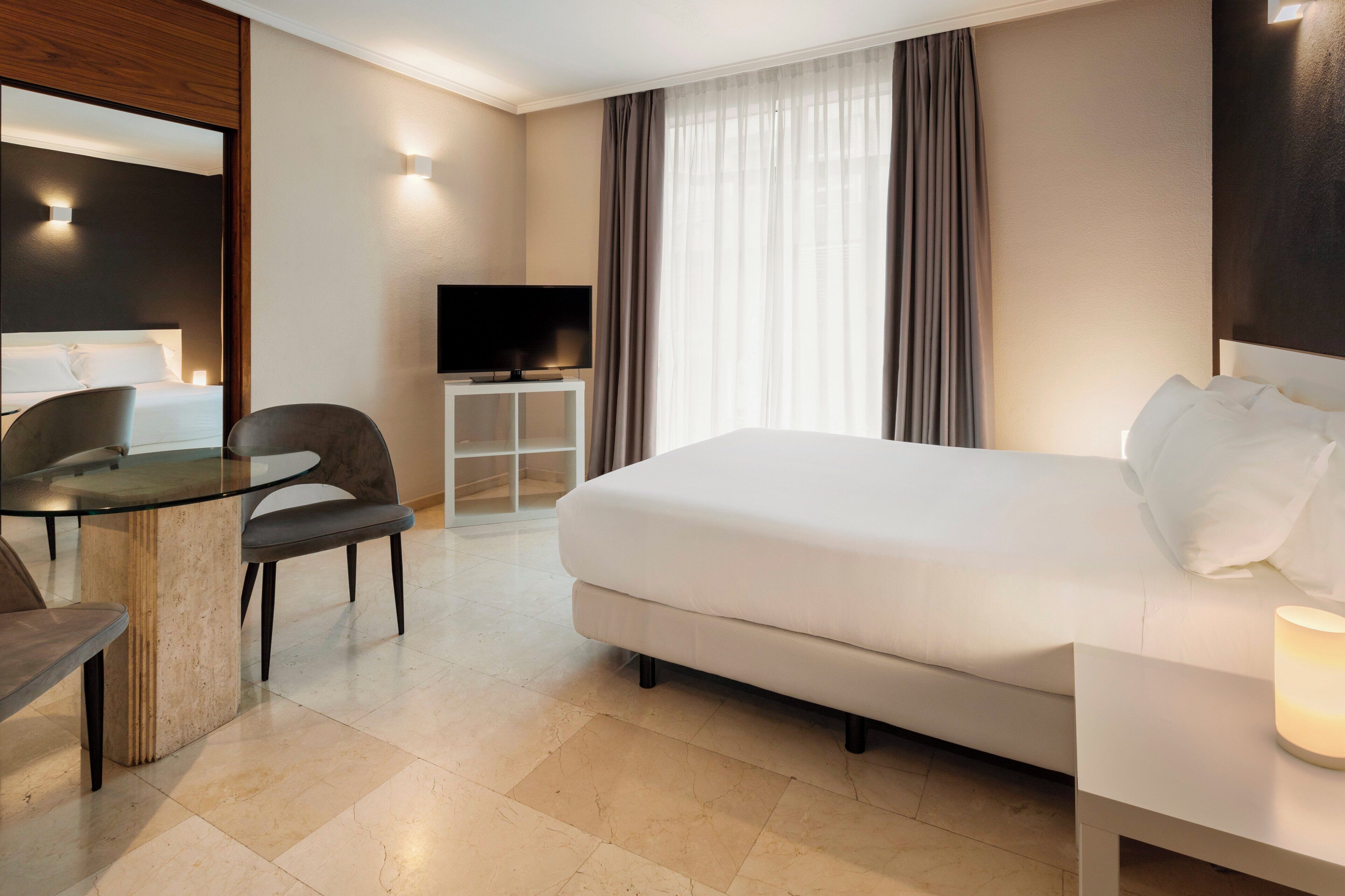 A hotel room with a large bed, a desk with a chair, a television on a white cabinet, and a window with curtains.