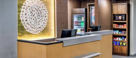 Uma área de recepção com balcão de madeira, um painel decorativo, um refrigerador com bebidas e um micro-ondas sobre uma prateleira com lanches.