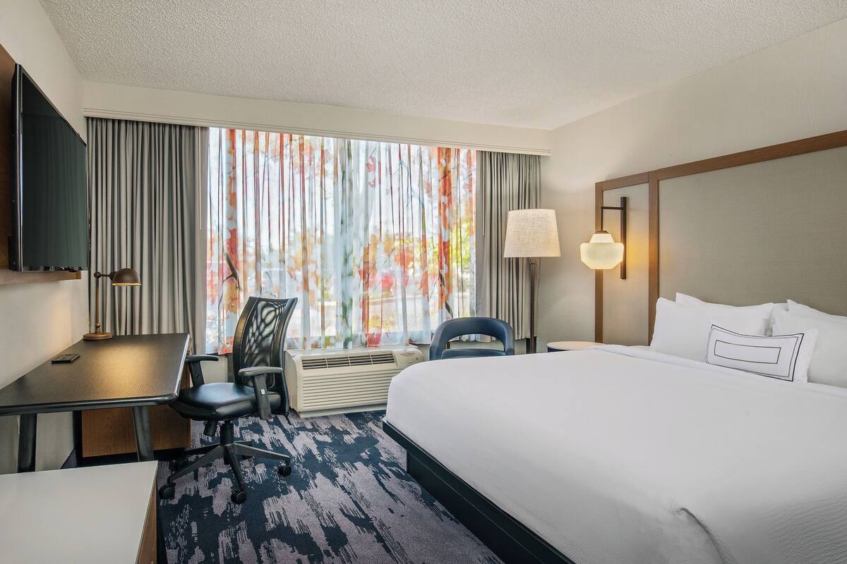 Une chambre d’hôtel équipée d’un lit, d’un bureau, d’une chaise, d’une télévision et d’une grande fenêtre avec des rideaux.