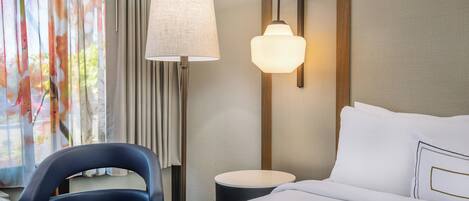 A hotel room with a bed, a desk, a chair, a lamp, and a wall-mounted light fixture.