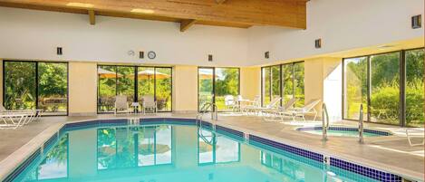 A swimming pool with clear blue water, surrounded by lounge chairs and umbrellas, with large windows offering views of greenery.