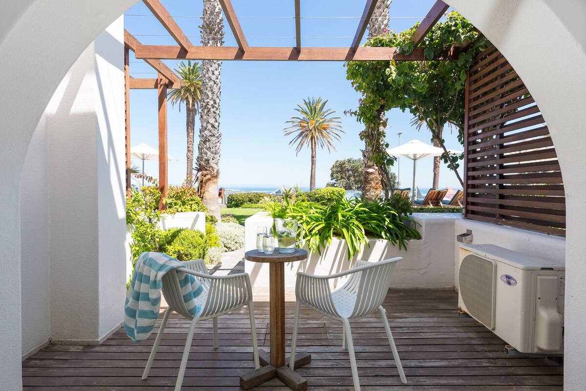 Eine Terrasse im Freien mit einem Holztisch und zwei weißen Stühlen, ein überdachter Sitzbereich mit Blick aufs Meer und eine Pergola.