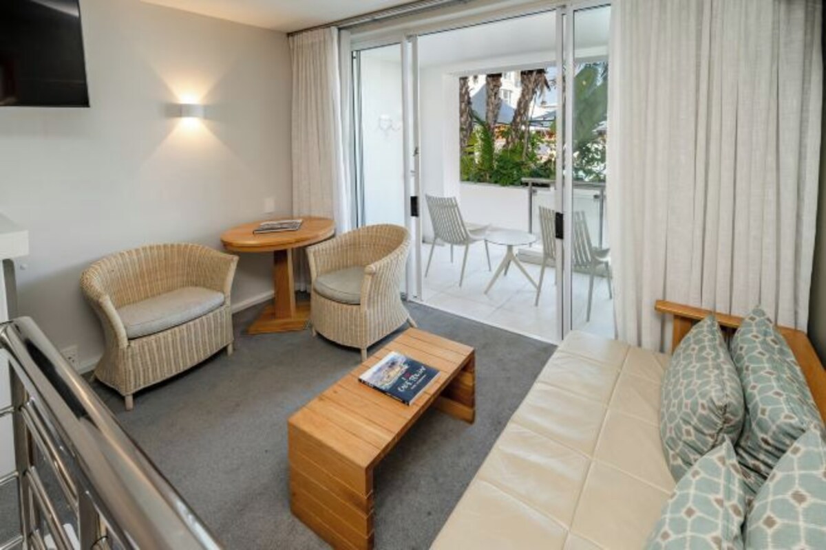A hotel room with a sofa, two armchairs, a coffee table, a TV, and a balcony with a table and chairs.