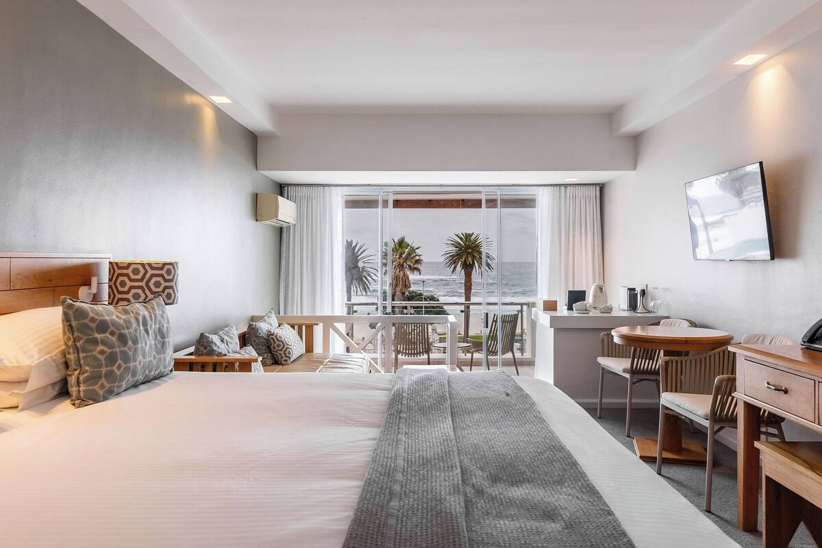 Ein Hotelzimmer mit Bett, Schreibtisch, Stuhl, kleinem Tisch und einem Balkon mit Meerblick.