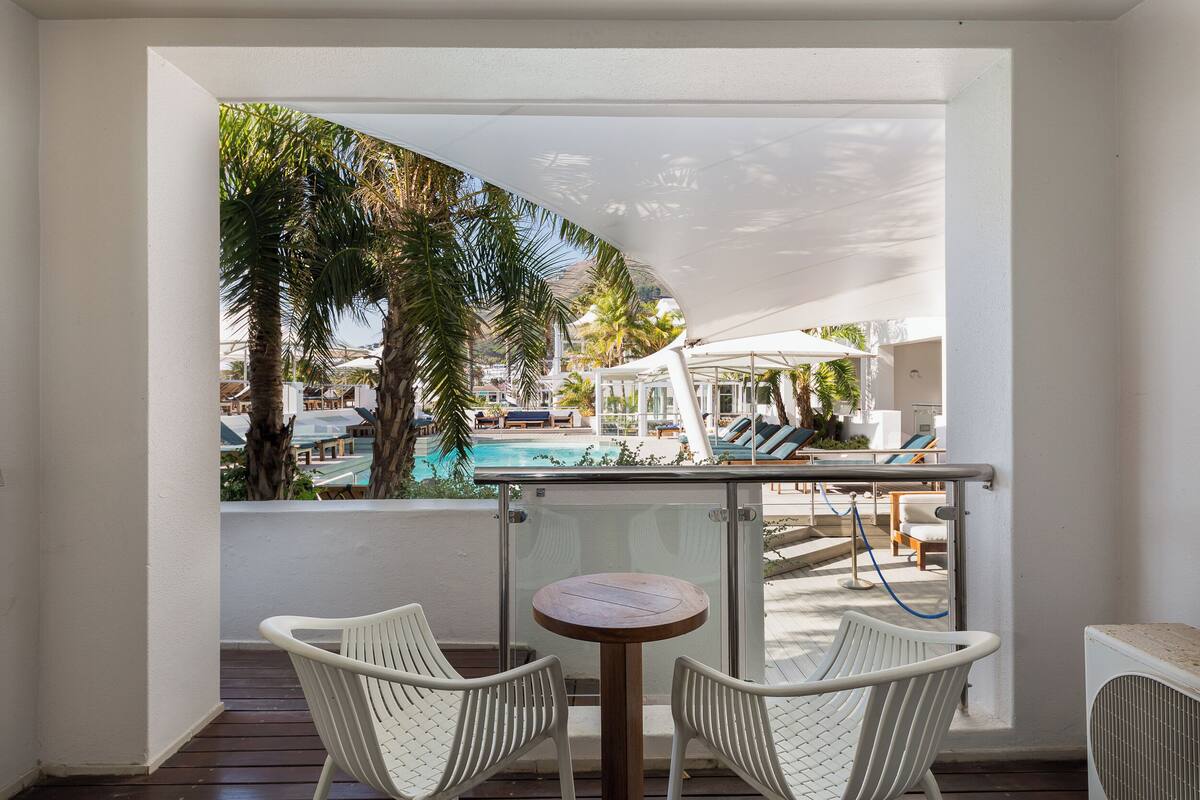 Ein Balkon mit zwei weißen Stühlen, einem kleinen runden Tisch und Blick auf einen Poolbereich mit Liegestühlen und Palmen.