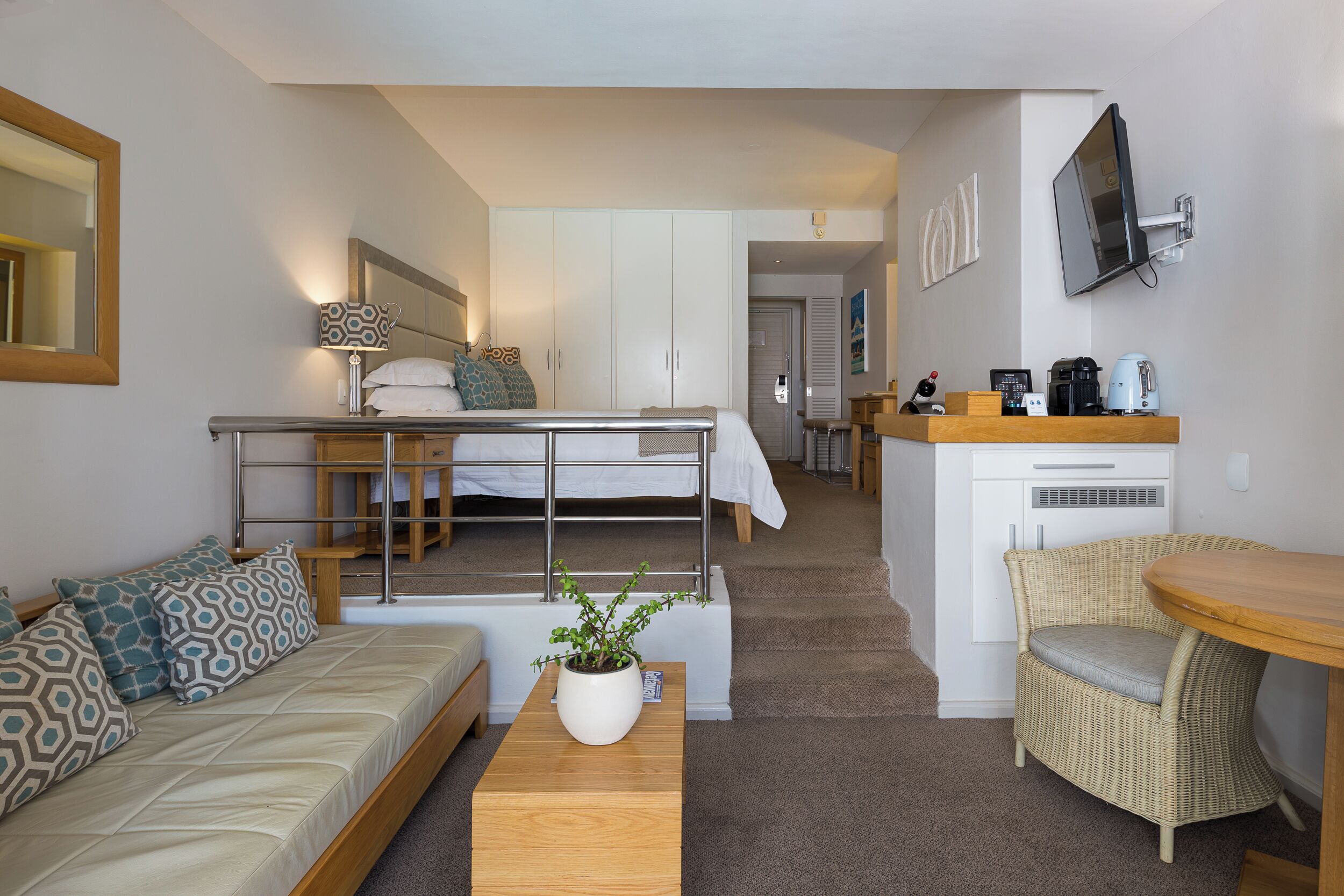 A compact hotel room with a sofa, a small table with a plant, a kitchenette, a dining area, and a separate bedroom with a bed and wardrobe.