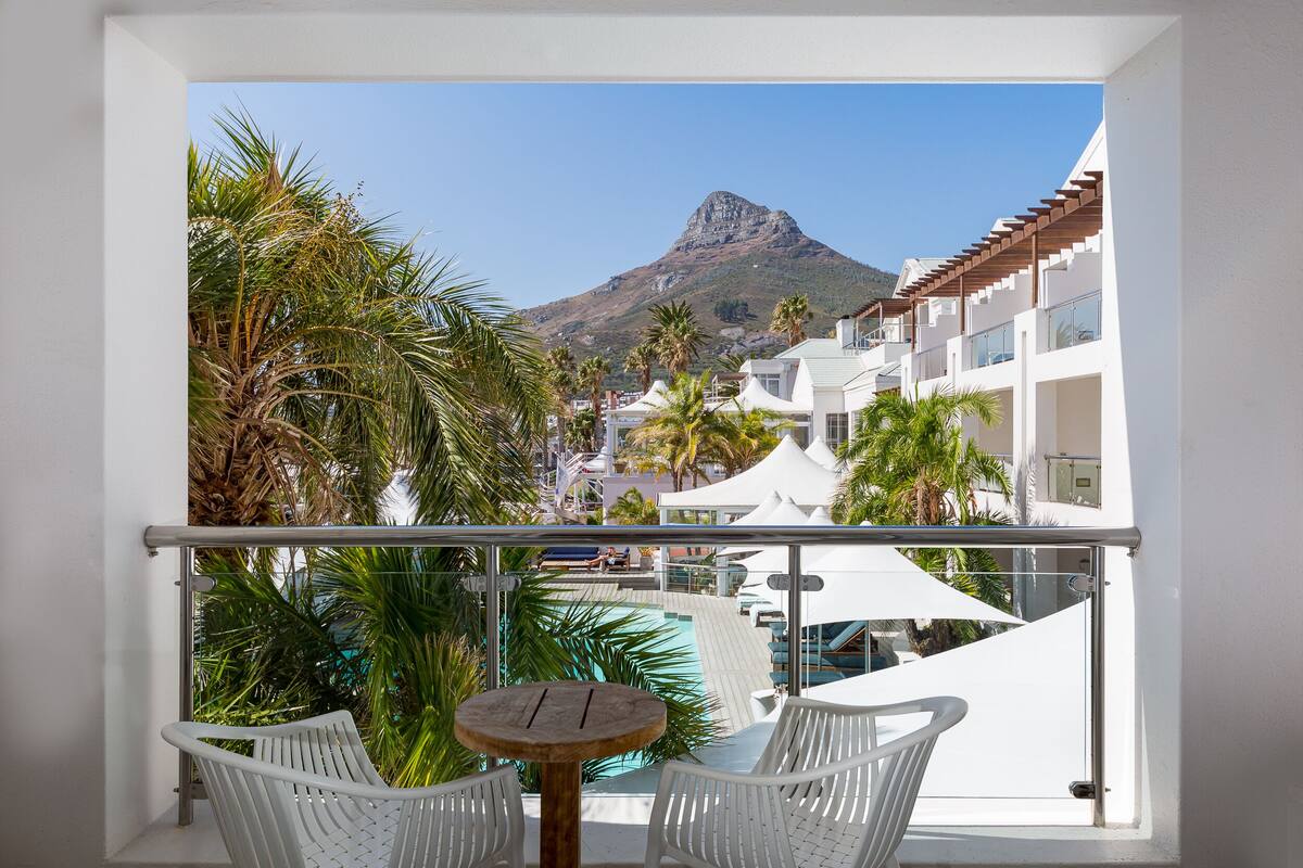 Ein Balkon mit weißen Korbstühlen und einem kleinen runden Tisch, mit Blick auf einen Poolbereich mit Palmen und einem Berg im Hintergrund.