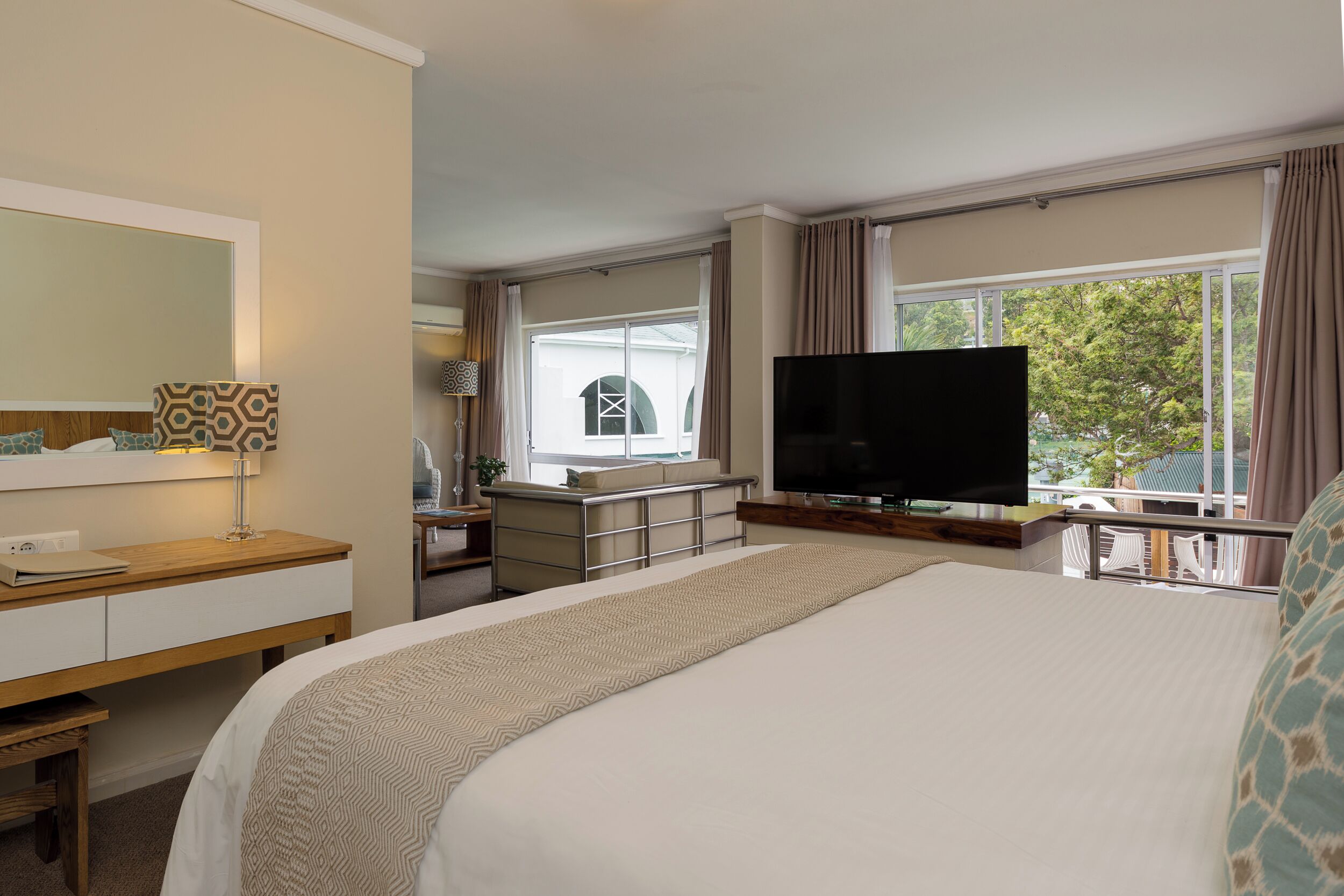 A hotel room with a large bed, a TV, a desk with a lamp, a dresser, and a window with curtains.