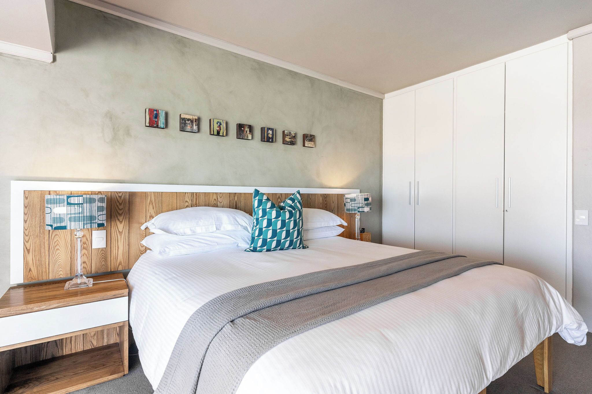 Una cama bien tendida con ropa de cama blanca y un cojín de diseño blanco y verde azulado, una mesita de noche con una lámpara y una pared con cuadros enmarcados.