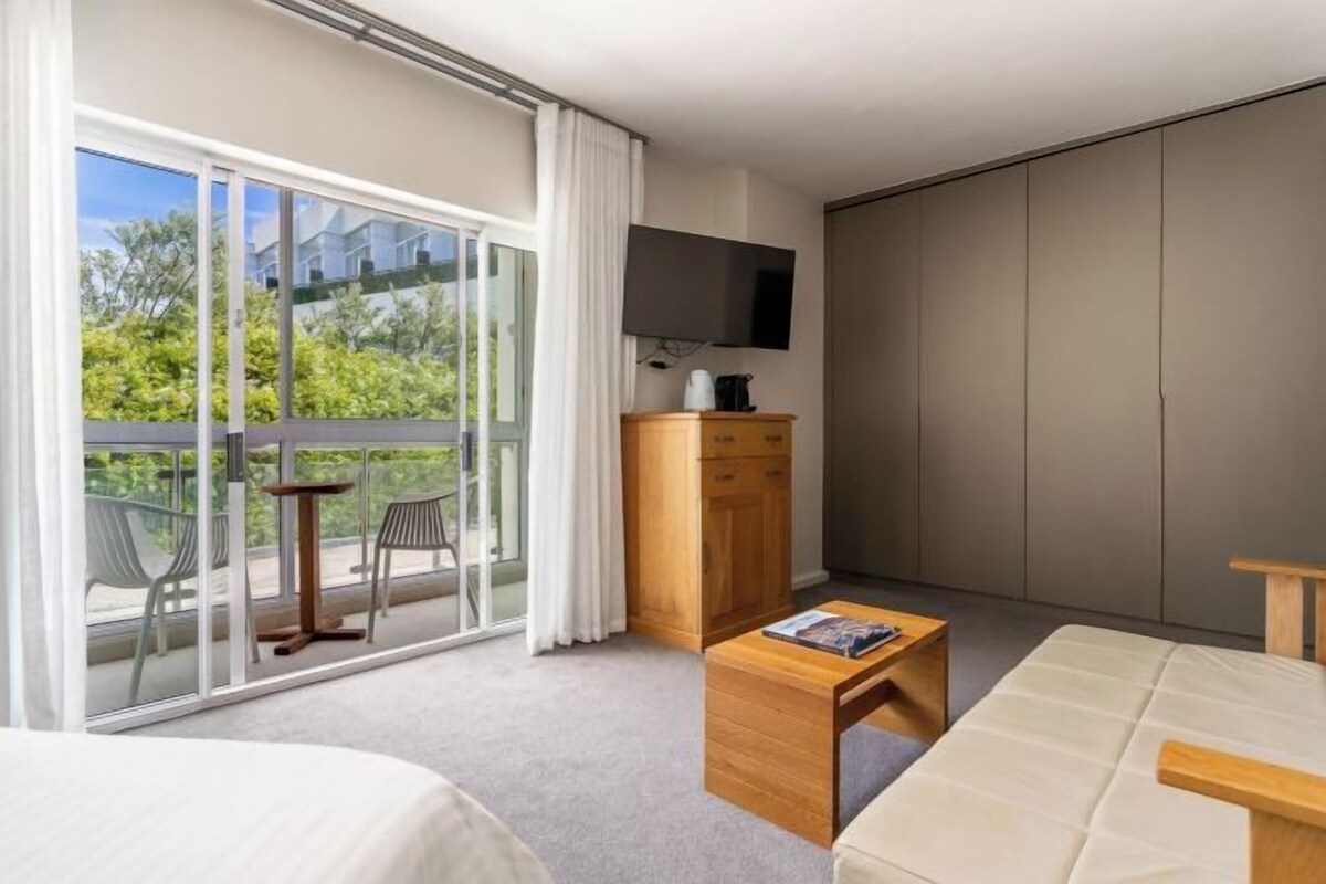 A hotel room with a bed, a wooden nightstand, a television, a sliding glass door to a balcony, and a view of greenery outside.