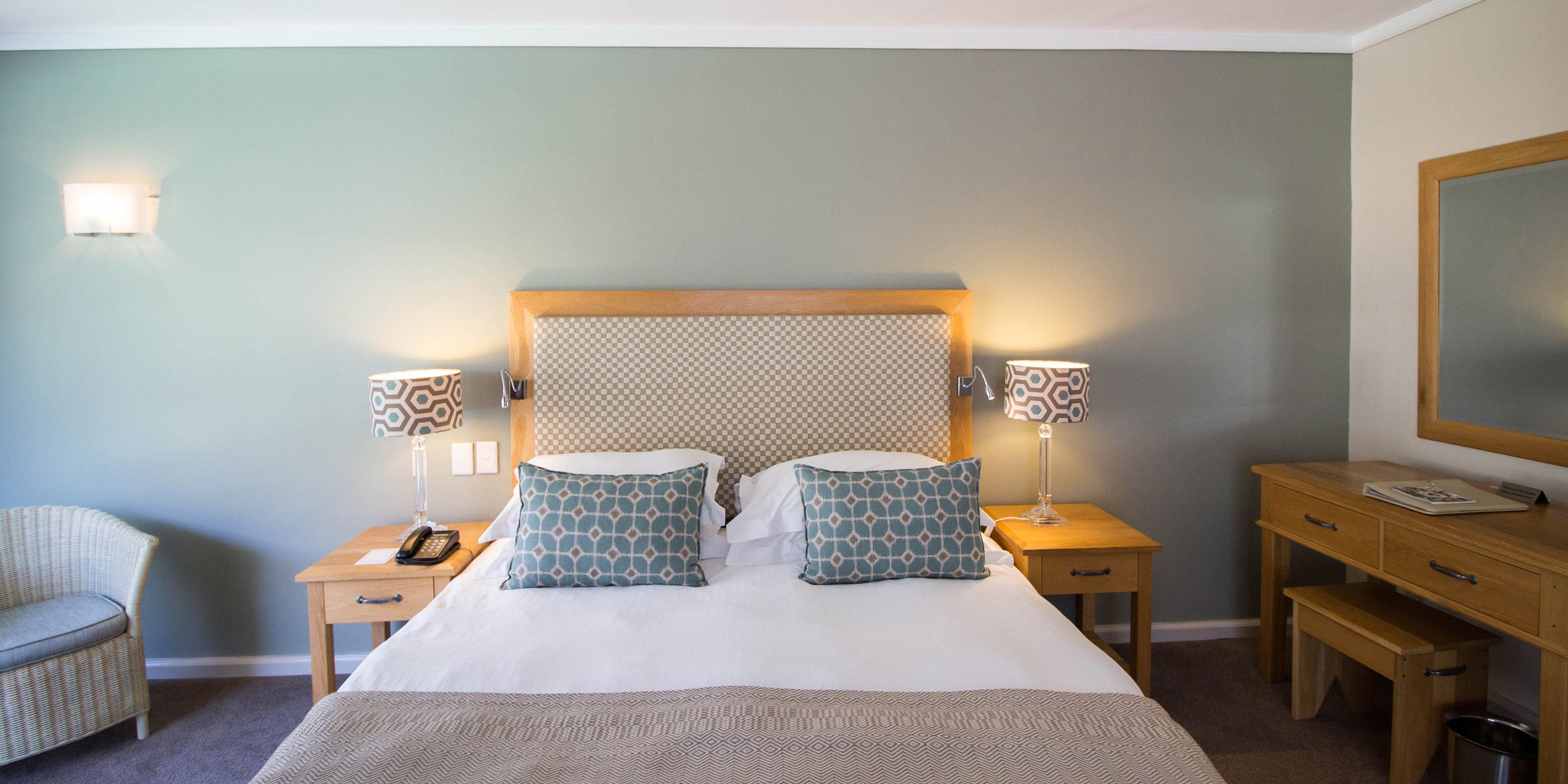 Habitación de hotel ordenada con una cama grande, dos mesitas de noche, una silla, una mesita auxiliar de madera y un espejo.