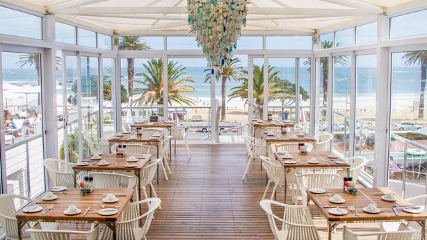 A covered dining area with tables and chairs, overlooking a beach with palm trees and the ocean. - The Bay Hotel (Cape Town)