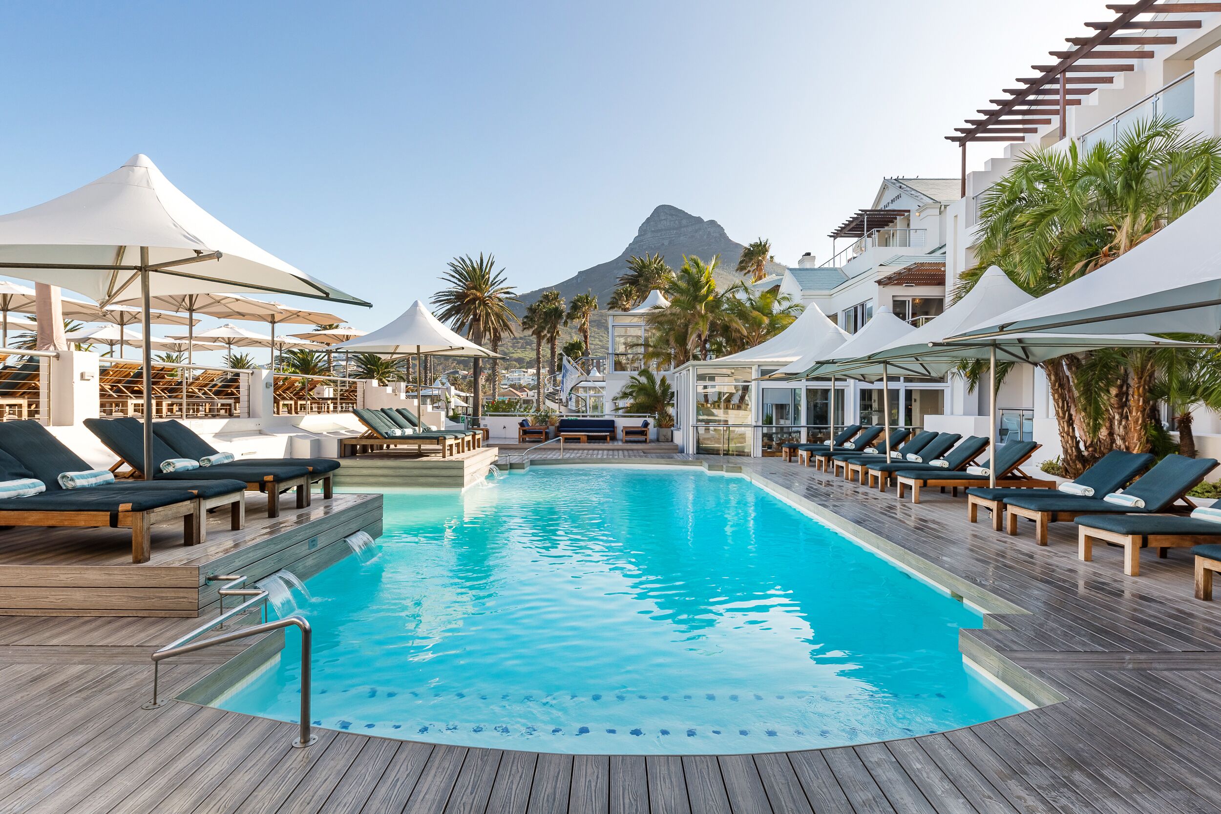 A pool area with lounge chairs and umbrellas, a wooden deck, and a mountain in the background.