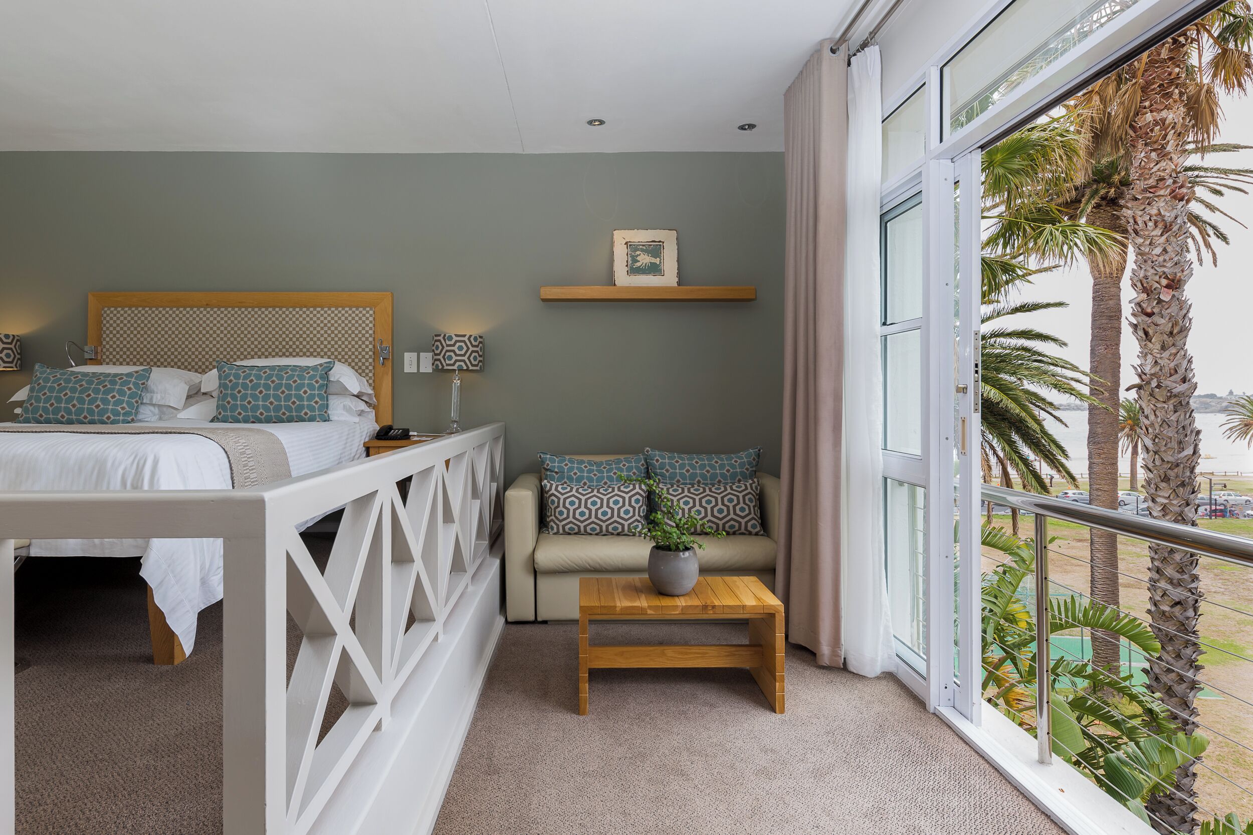 A bedroom with a bed, a sofa, a small table, and a window with a view of palm trees.