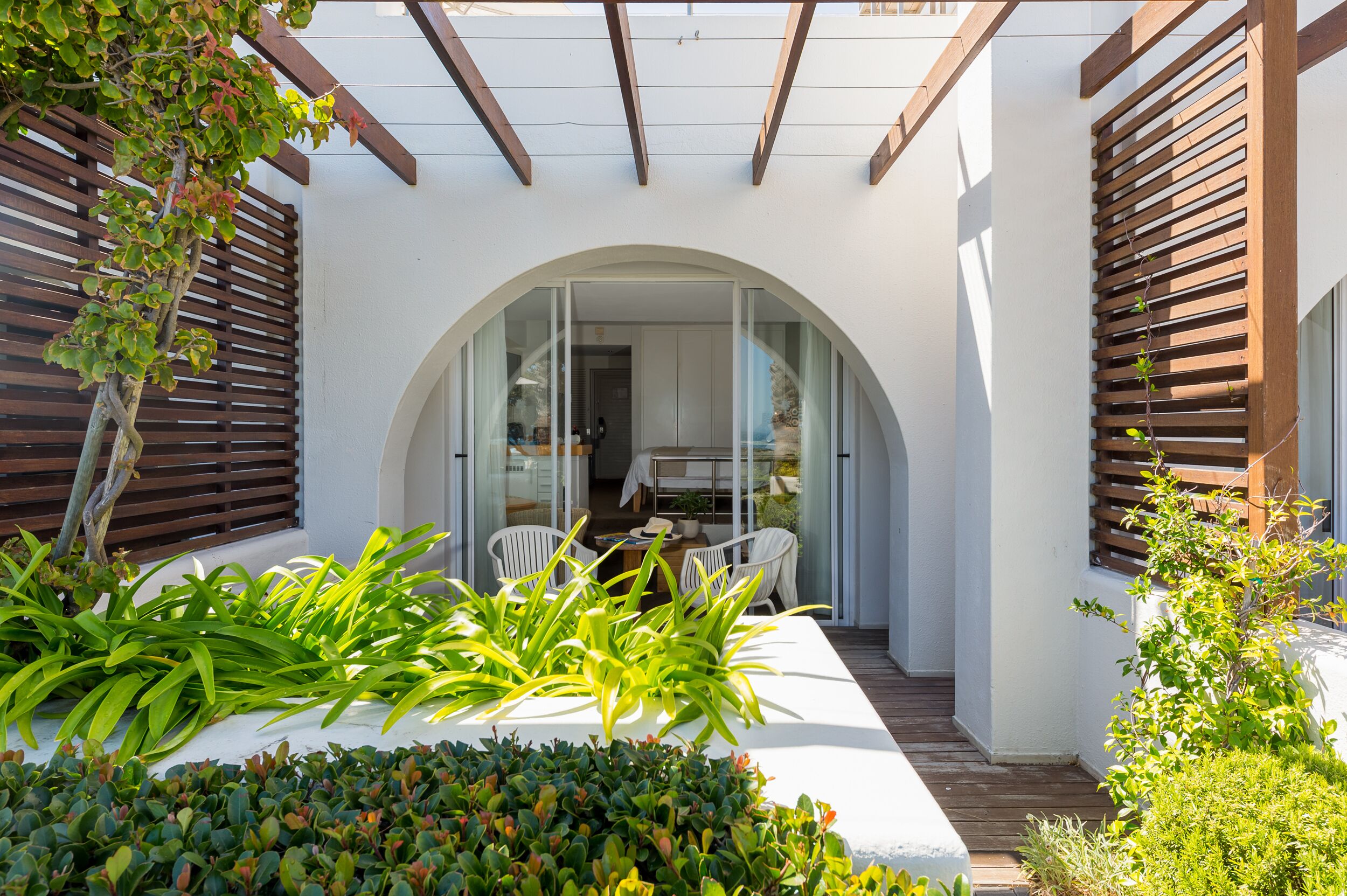 A covered patio with a white table and chairs, surrounded by greenery and wooden slats.