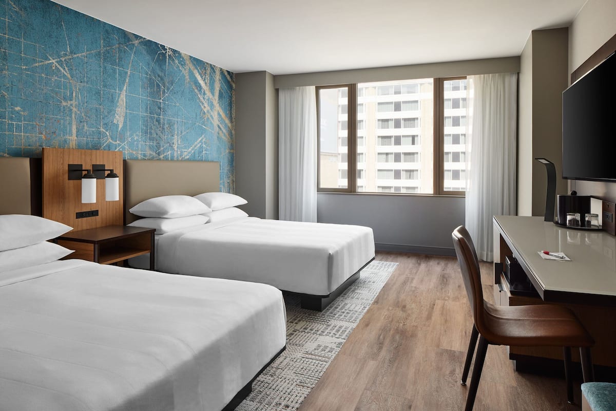 A hotel room with two beds, a desk, a chair, a TV, and a large window with curtains.