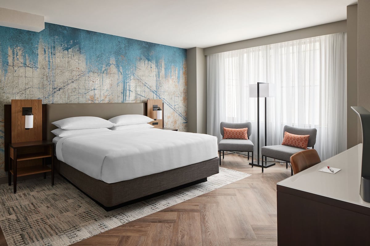 A modern hotel room with a large bed, a desk, two chairs, and a large window with sheer curtains.