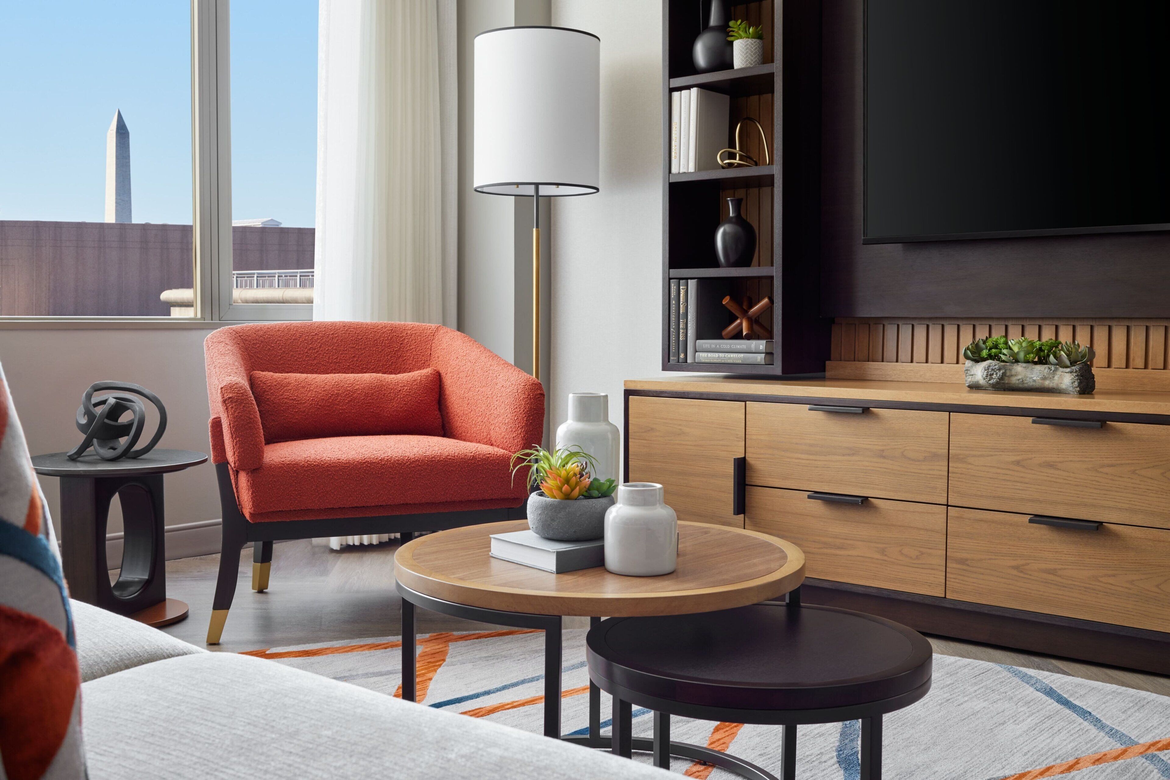 A modern living room with a red armchair, a round wooden table, a floor lamp, a bookshelf, and a TV mounted on the wall.