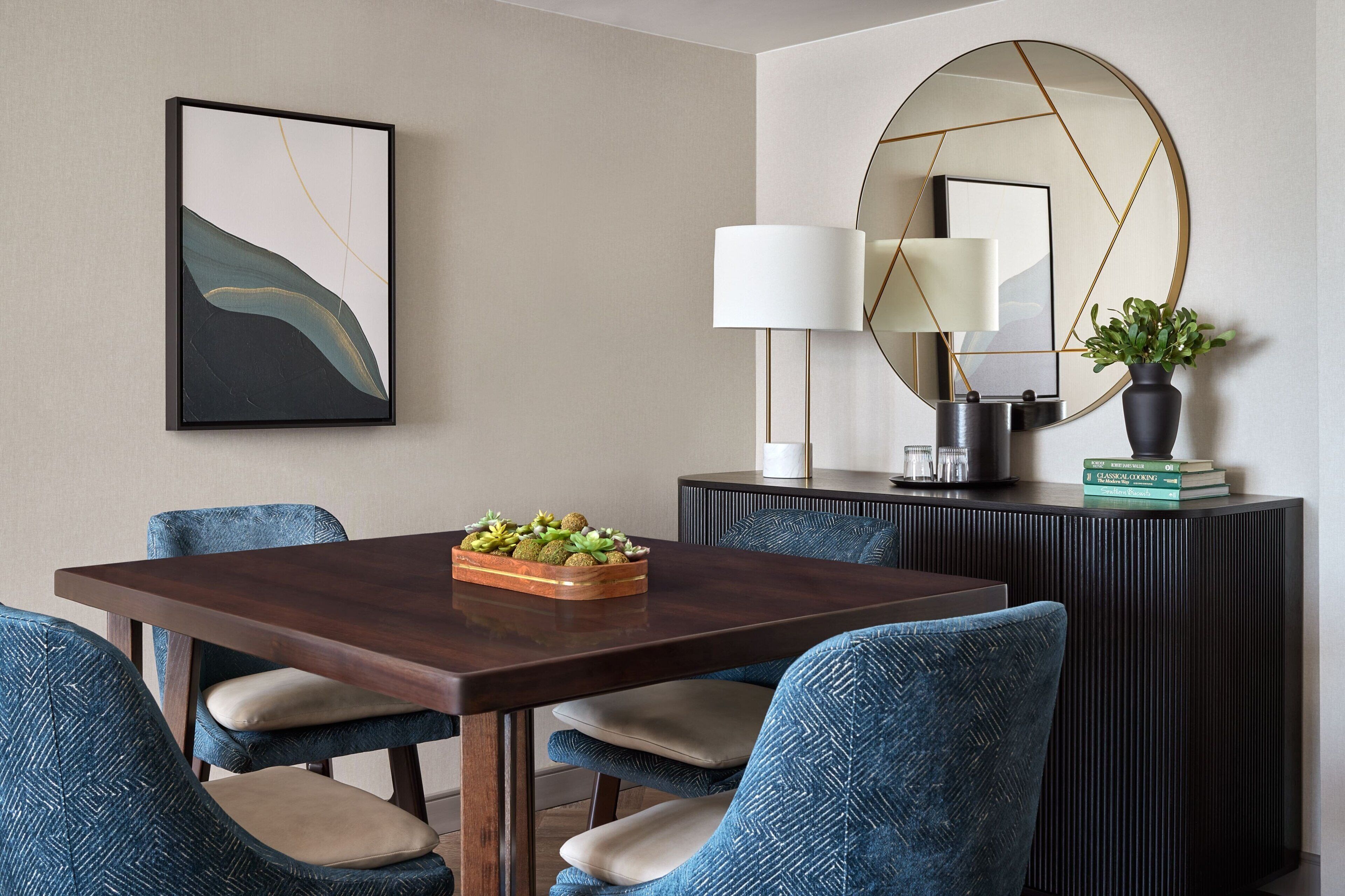 A dining area with a rectangular table, four chairs, a decorative bowl, a wall-mounted mirror, a lamp, and a potted plant.