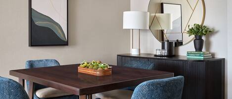 A dining area with a rectangular table, four chairs, a decorative bowl, a wall-mounted mirror, a lamp, and a potted plant.