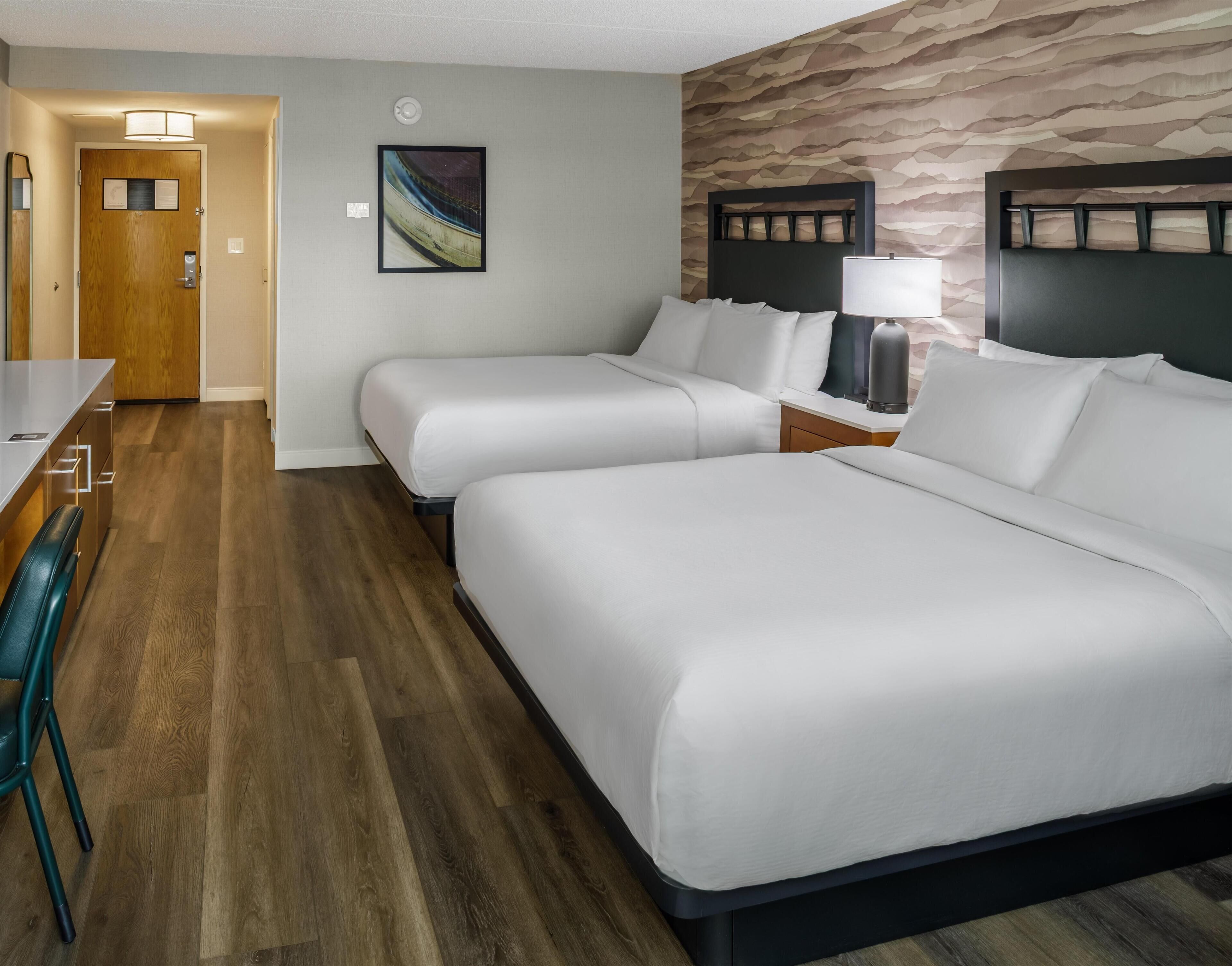 A hotel room with two beds, a desk, a chair, a lamp, and a wall with a textured pattern.