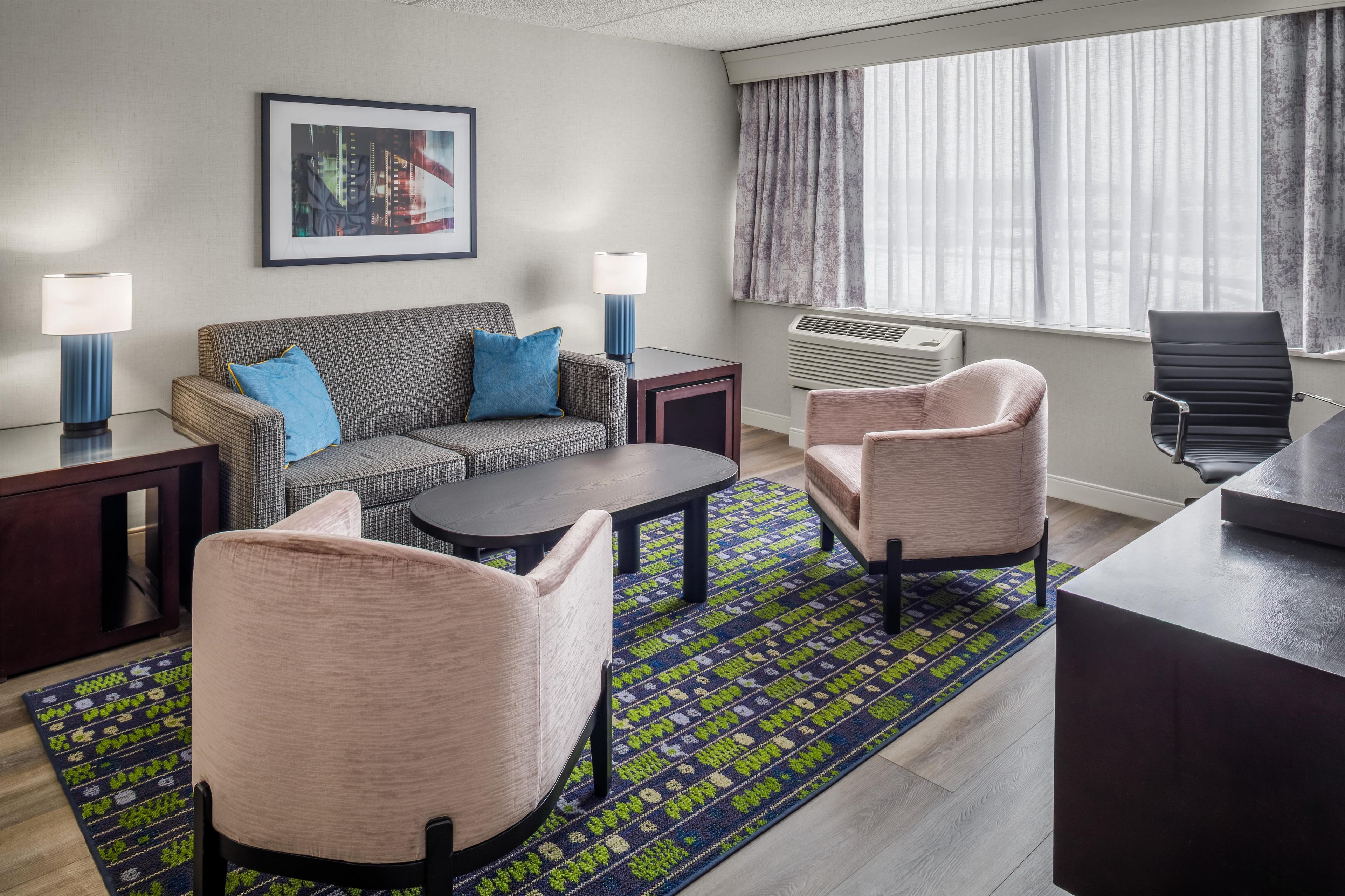 Une chambre d’hôtel avec un canapé, deux fauteuils, une table basse, un bureau et une grande fenêtre avec des rideaux.