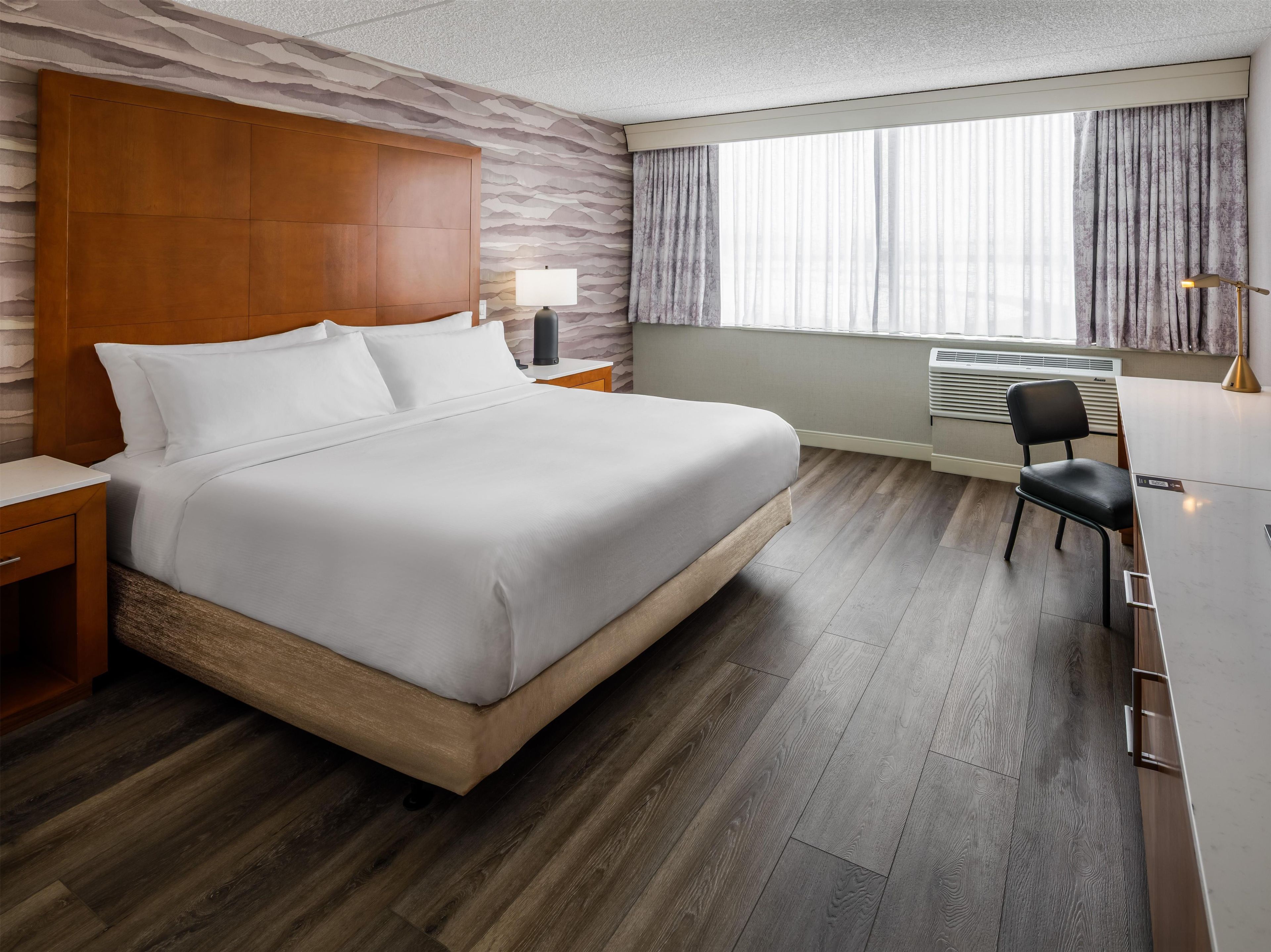 A hotel room with a bed, bedside table, lamp, chair, and a window with curtains.