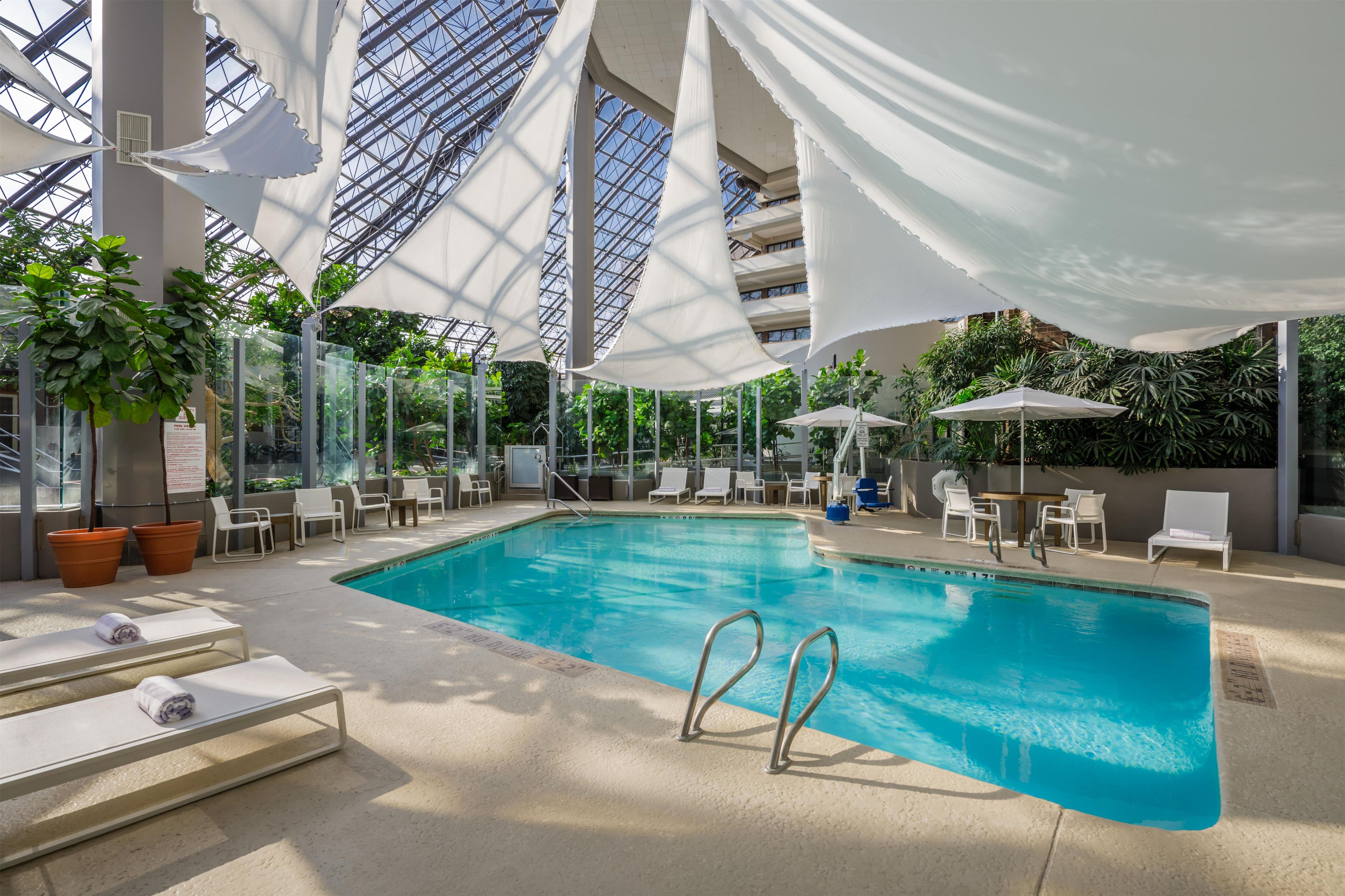An indoor swimming pool with clear glass ceilings, surrounded by lounge chairs and potted plants.