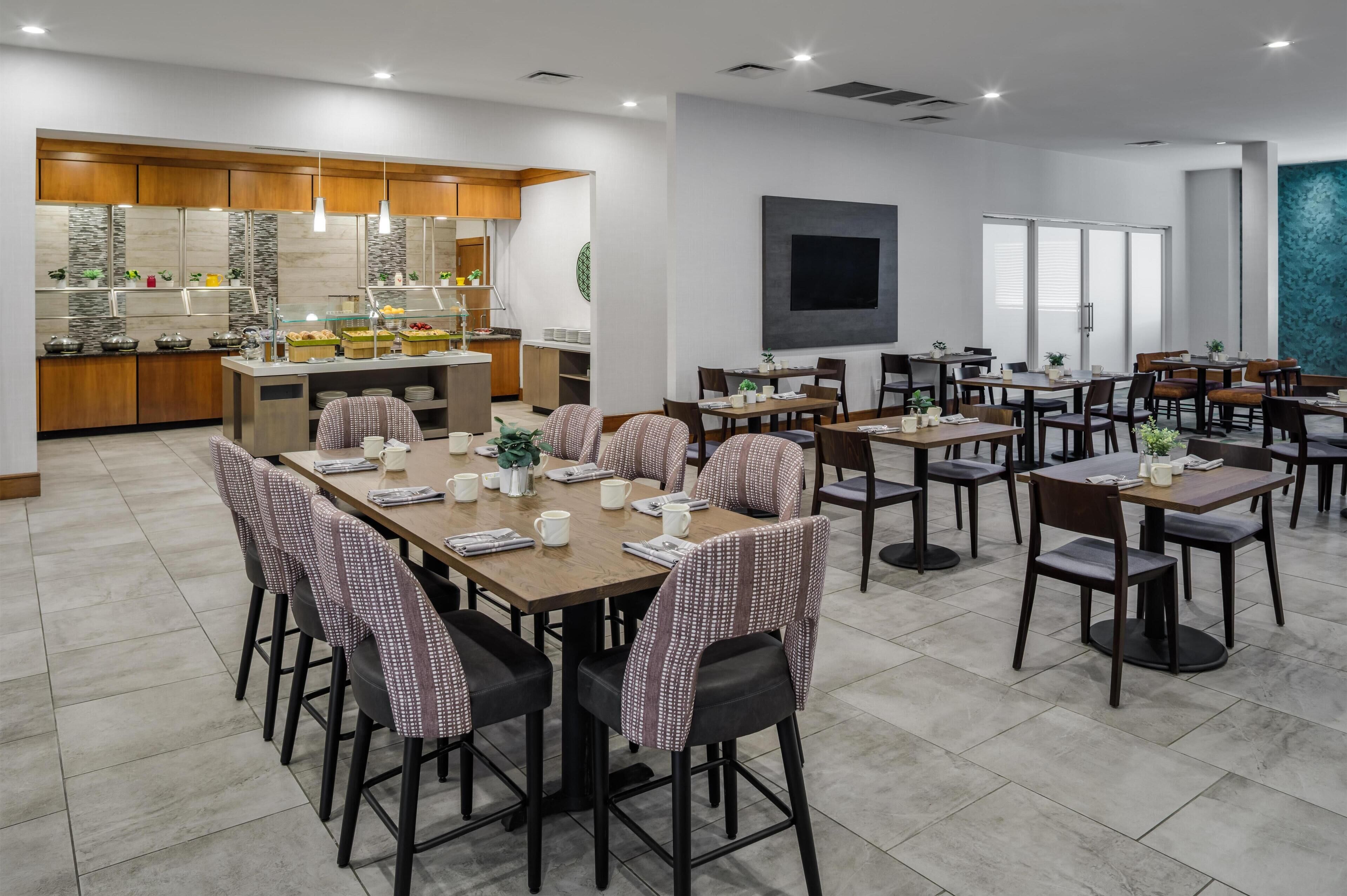 Un'area ristorazione d'hotel con diversi tavoli e sedie, una zona cucina con buffet e un grande schermo piatto a parete.
