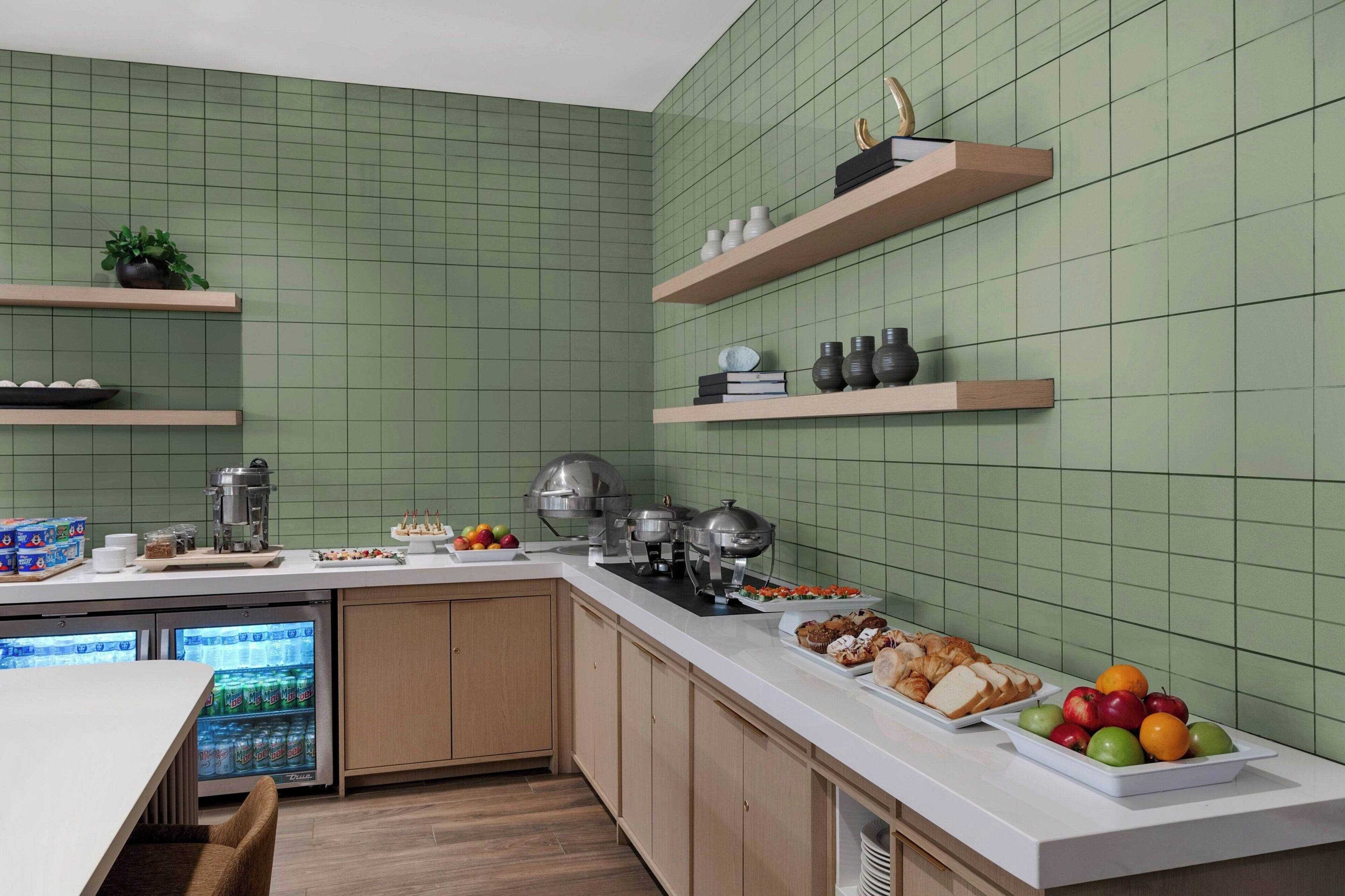 A kitchen with green tiled walls, wooden cabinets, a white countertop, and various food items and kitchen appliances.