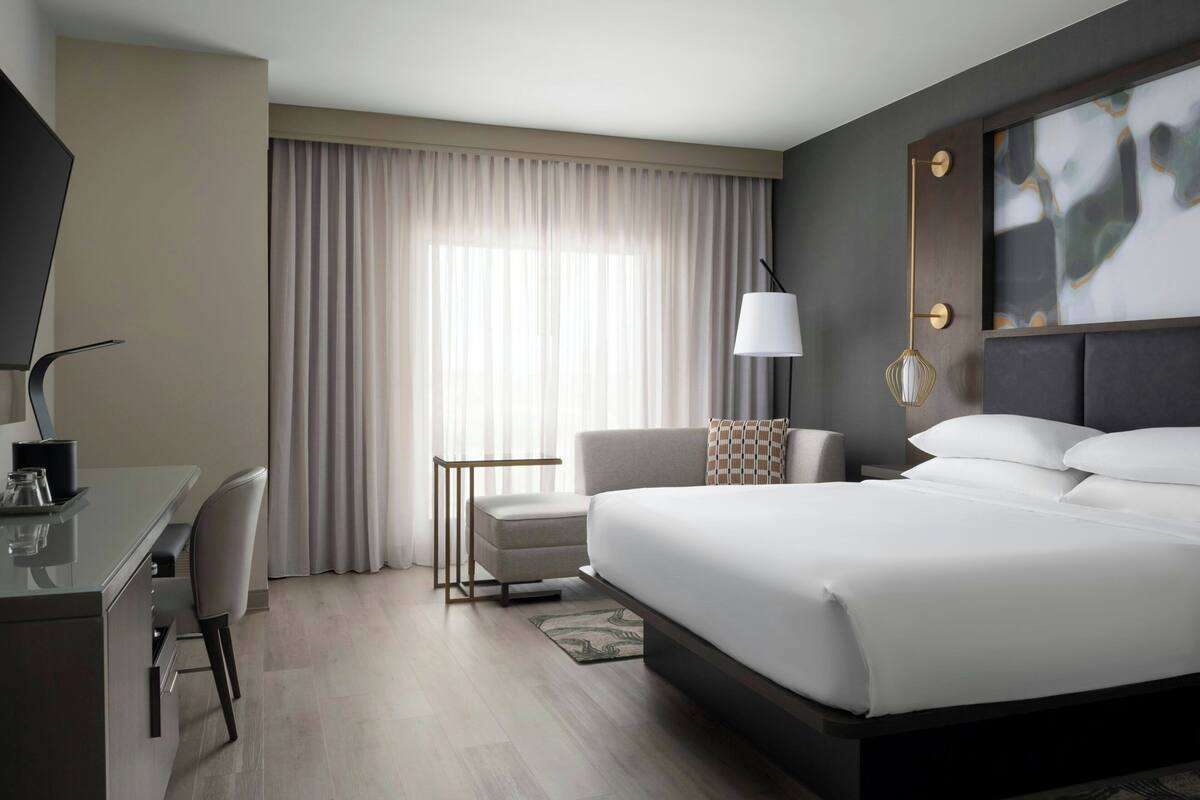 A modern hotel room with a large bed, a desk, a chair, and a window with curtains.