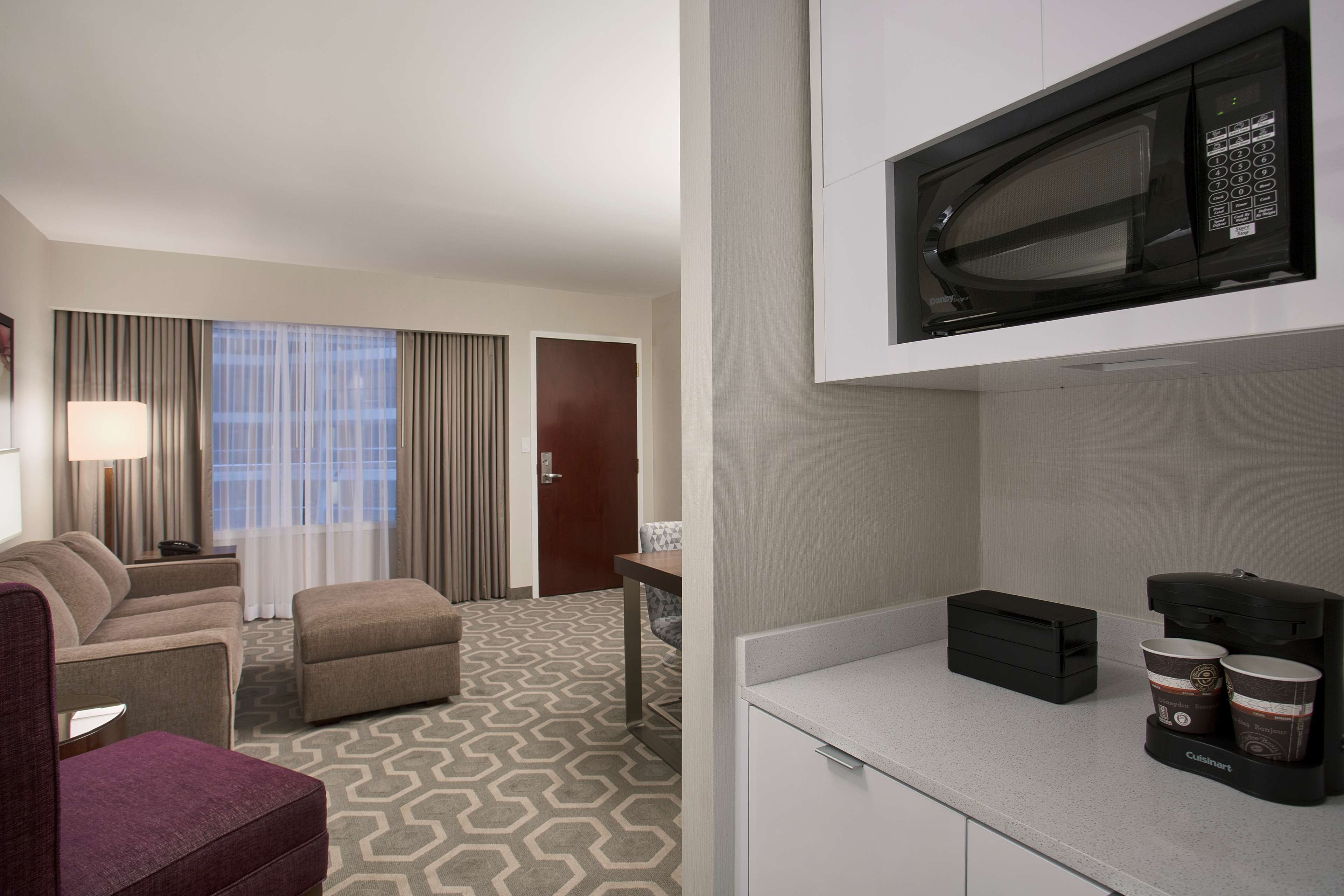A hotel room with a sofa, armchair, and ottoman, a kitchenette with a microwave, and a coffee maker.