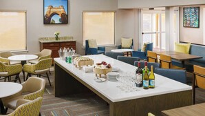 A hotel dining area with round tables, blue and yellow chairs, a central island with food and drinks, and large windows with blinds. - Residence Inn by Marriott Hartford/Windsor (Windsor)