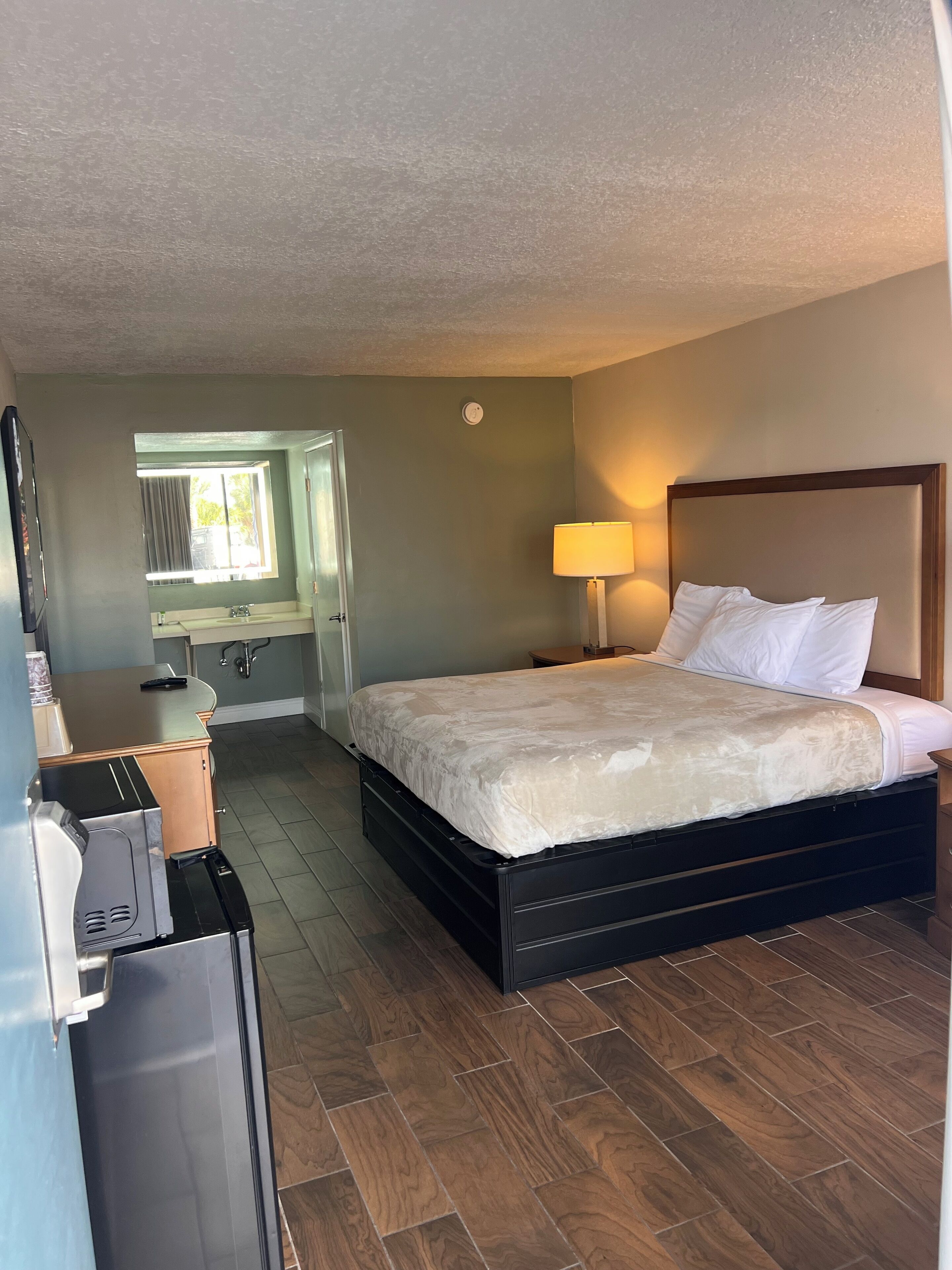 Habitación de hotel con cama, lámpara de noche, espejo, ventana y kitchenette.