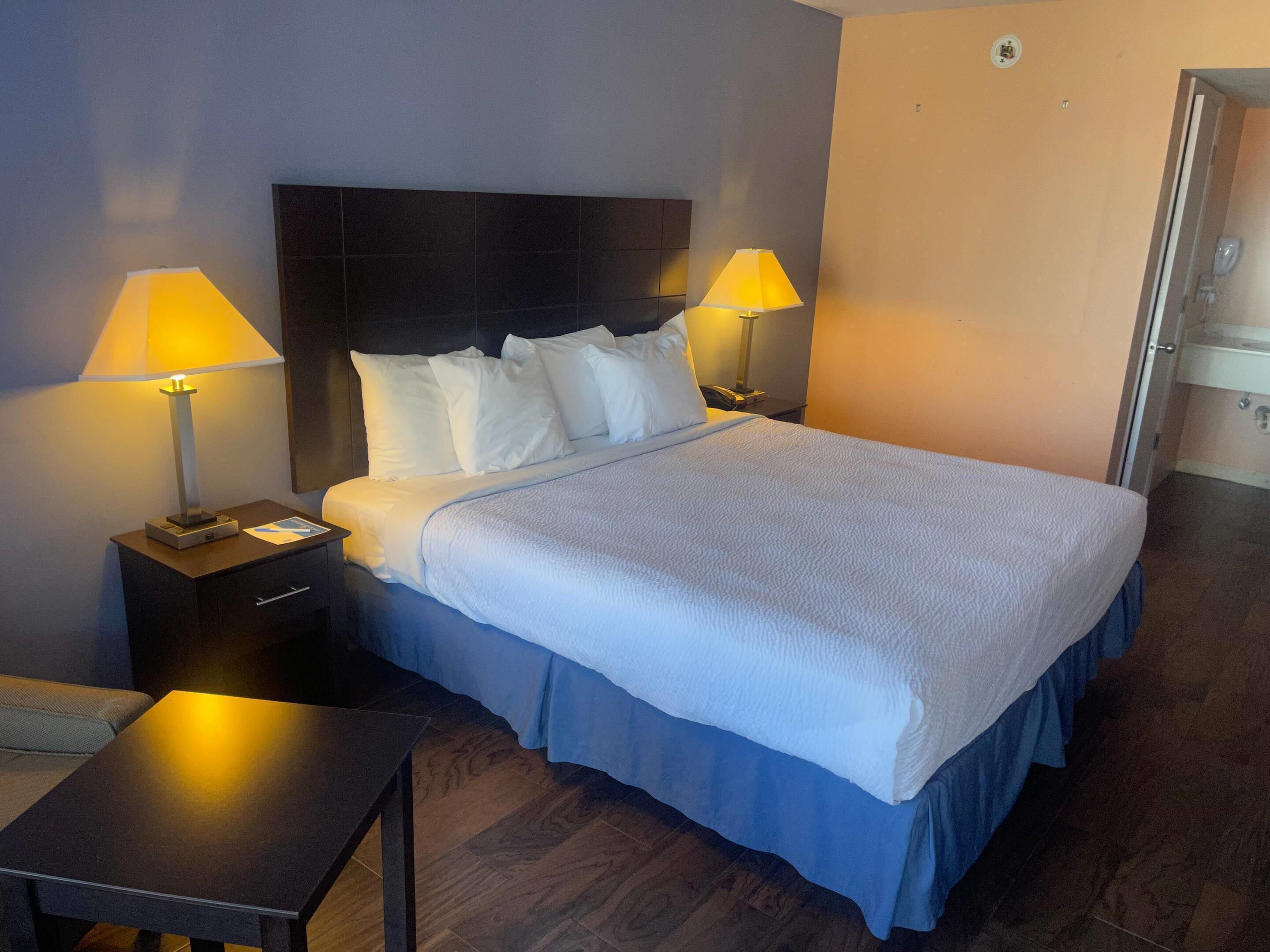 Ein Hotelzimmer mit einem großen Bett, zwei Nachttischlampen, einem Nachttisch, einem kleinen Tisch und einer Tür zu einem weiteren Raum.