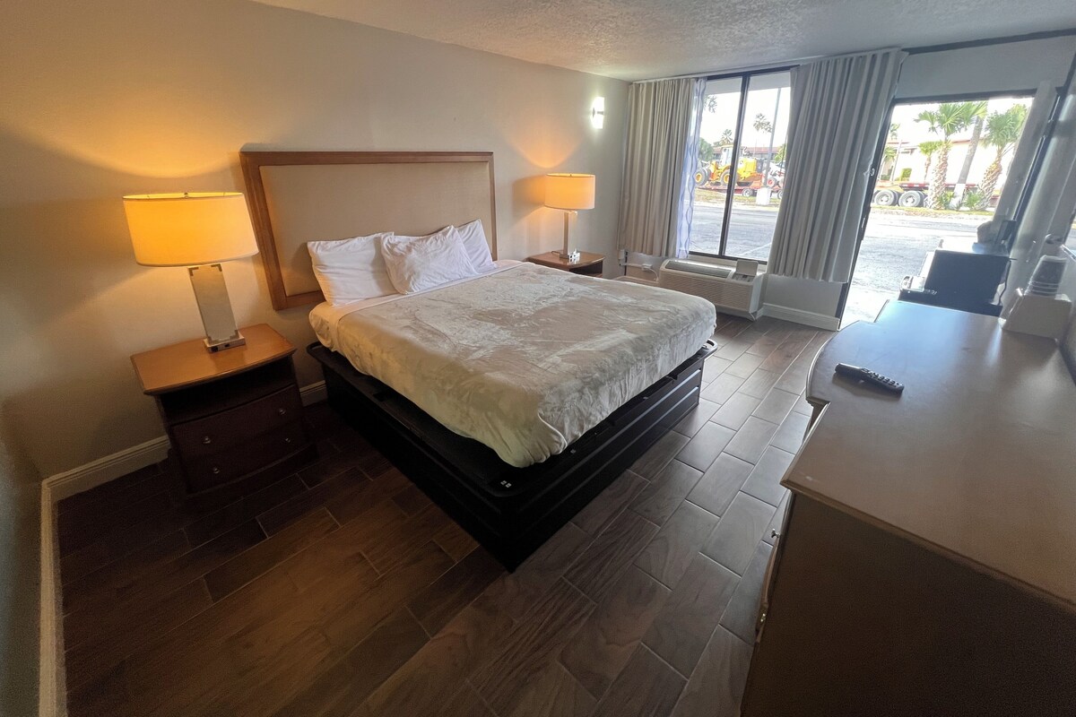 A hotel room with a large bed, two bedside lamps, a wooden nightstand, a window with curtains, and a view of the outside.