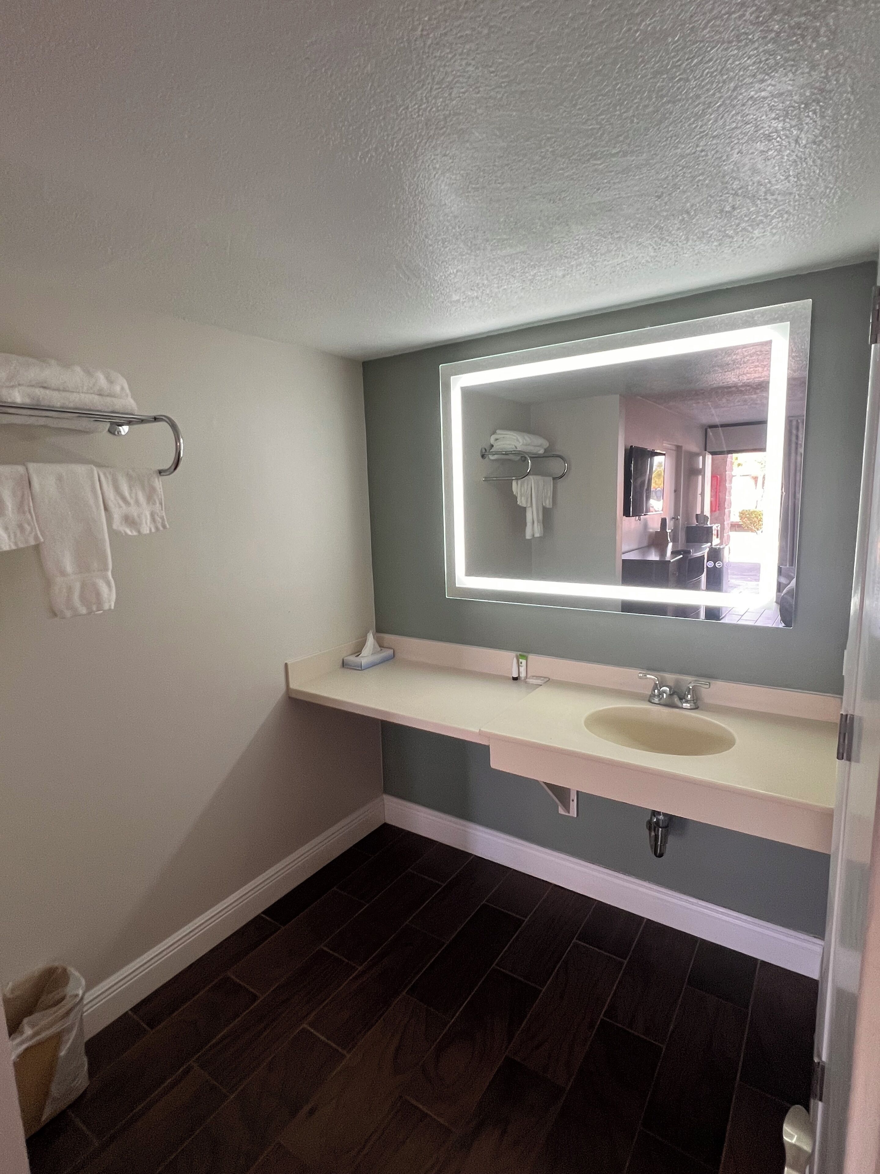 A bathroom with a white sink, a mirror with lights, a towel rack, and dark wooden flooring.