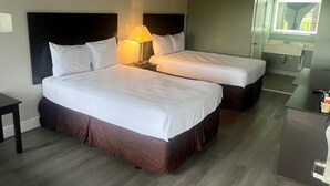 A hotel room with two beds, a nightstand with a lamp, a small table, a desk, and a mirror above the desk.